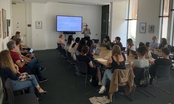A classroom of summer school students sit around a large table, facing a screen showing text